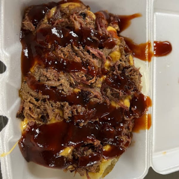 A Bama Boys BBQ loaded baked potato with pulled pork, cheese, and barbecue sauce in a takeout container.